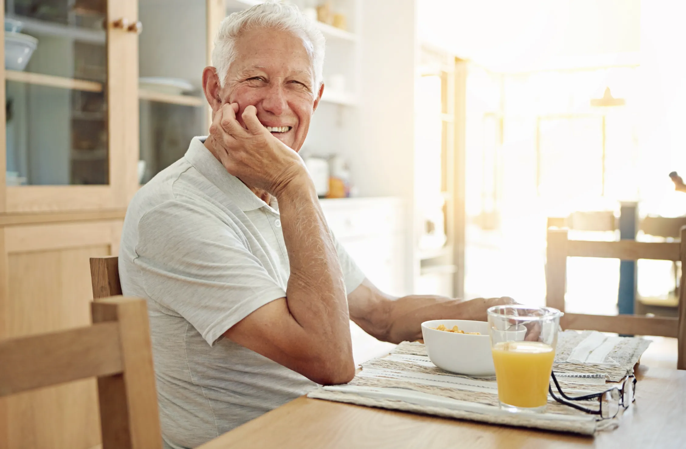 Hombre mayor sonriendo mientras desayuna con cereal y jugo de naranja en casa.