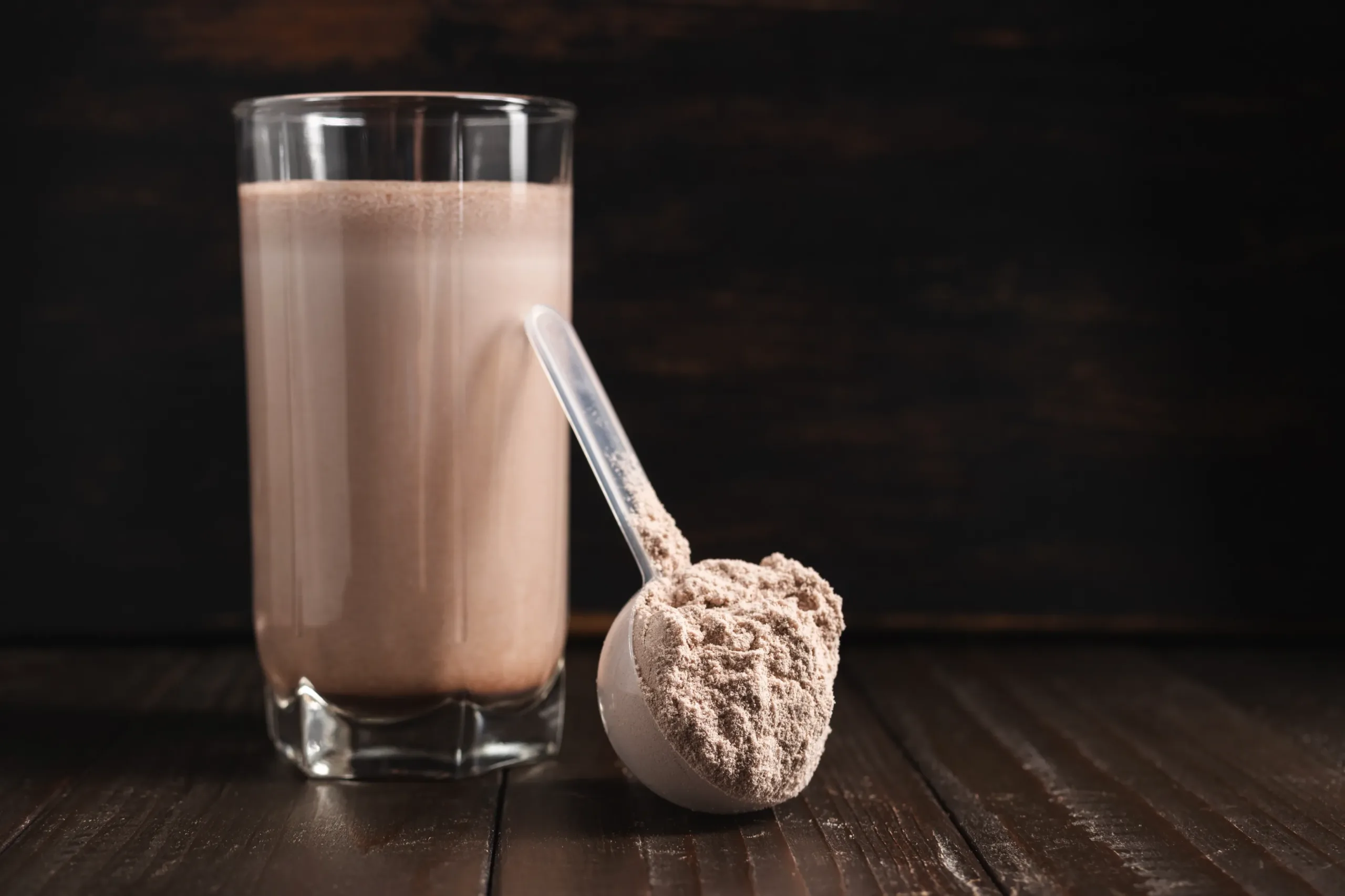 Vaso con batido de proteína de chocolate junto a una cucharada de polvo proteico