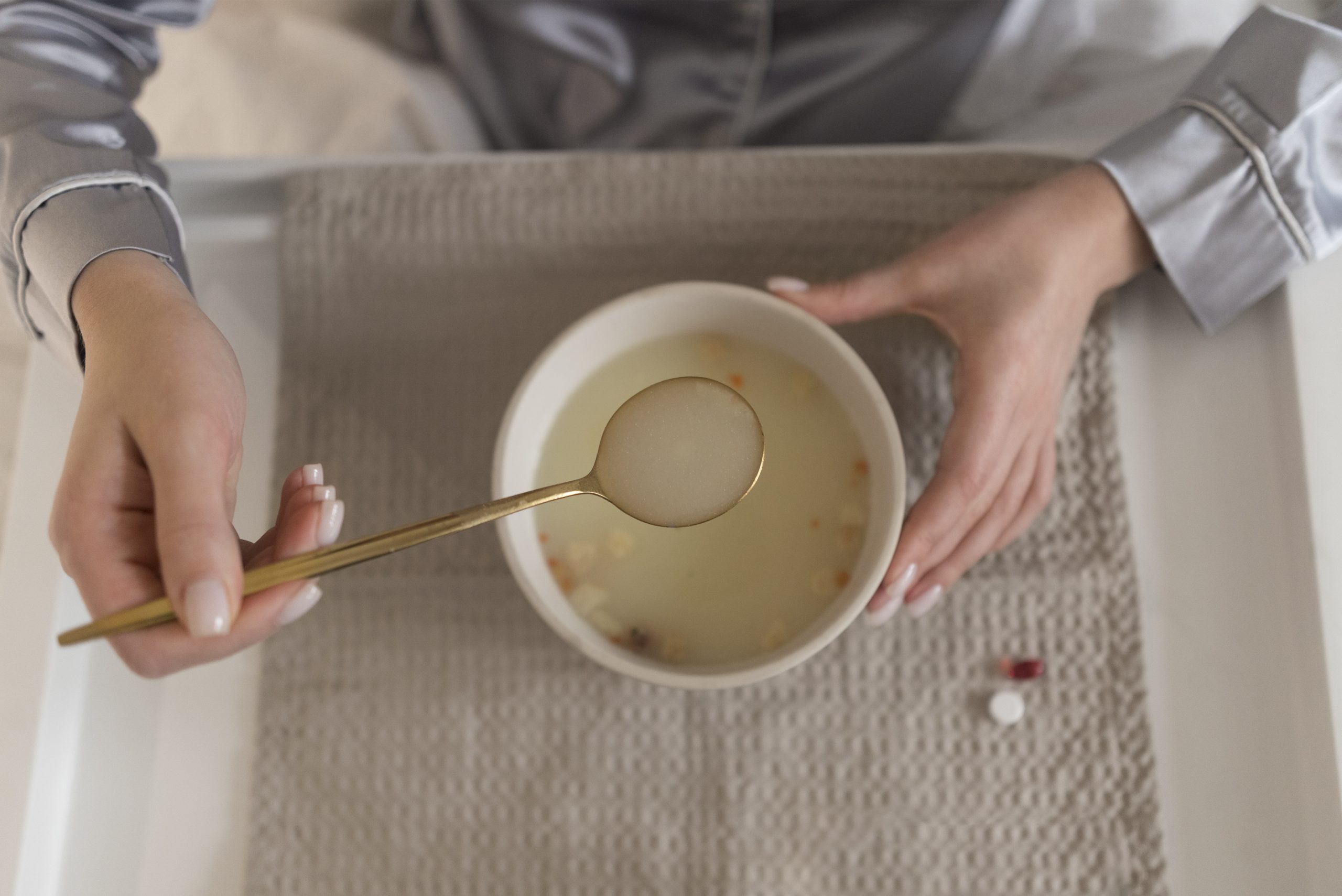 ujer con pijama sosteniendo una cuchara de sopa caliente en una bandeja con medicamentos al lado