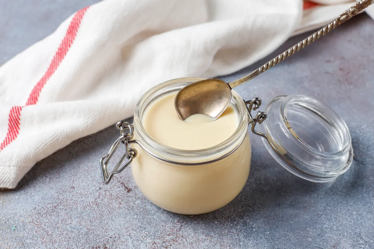 Frasco de vidrio abierto con leche condensada y una cuchara metálica encima